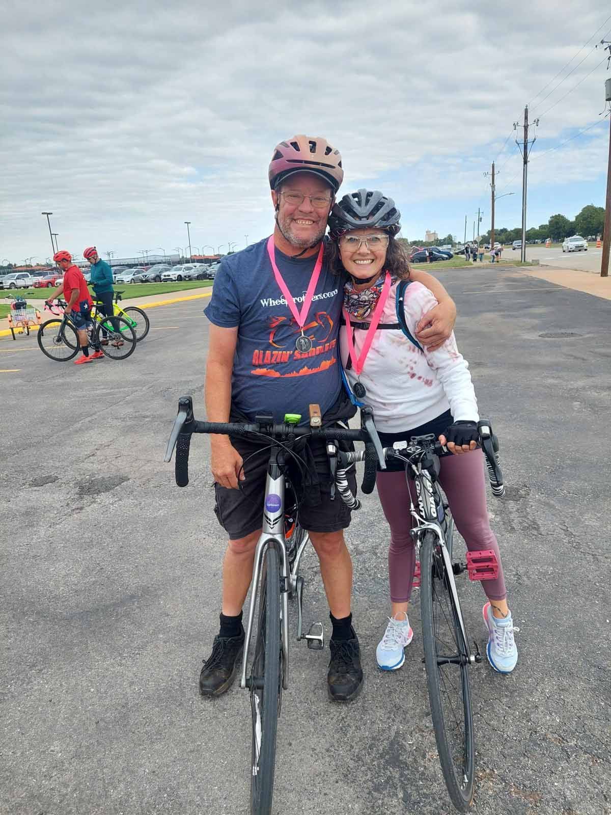 Cyclists pose together, smiling, after a bike ride. They wear helmets and pink ribbons. Outdoors, asphalt, cloudy sky.