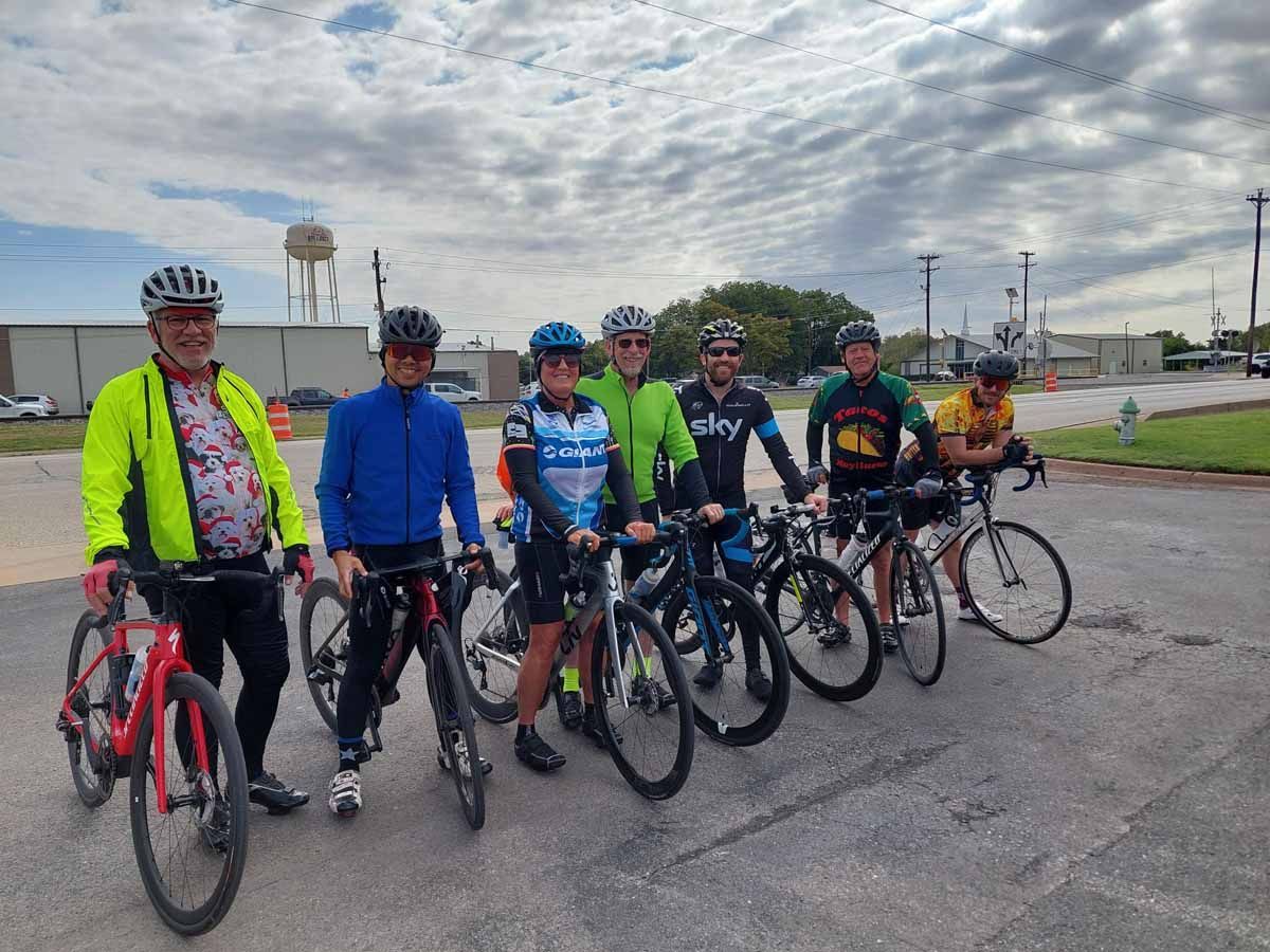 Cyclists in helmets and jerseys, standing with their bikes on a road. Cloudy sky, water tower in background.