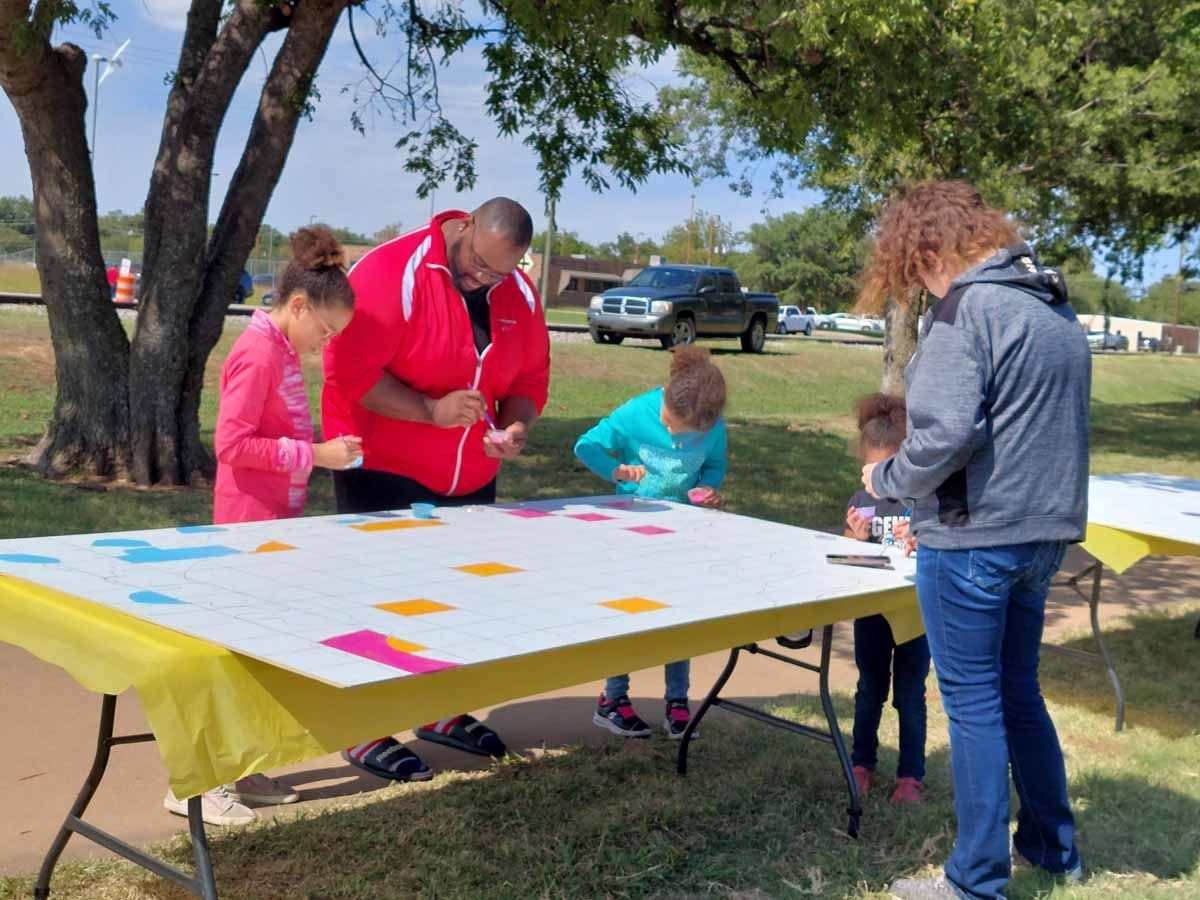 People crafting on a large table outdoors, applying colorful shapes.