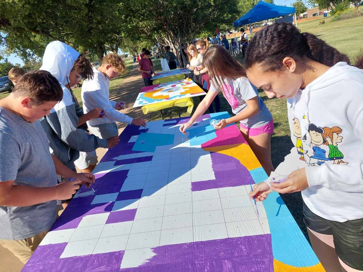 Students painting a large, colorful mural on tables outdoors.