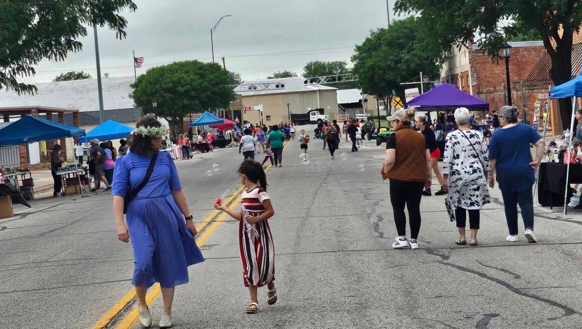 People walking along a closed street with vendors and tents. Cloudy day, blue and white colors are prominent.