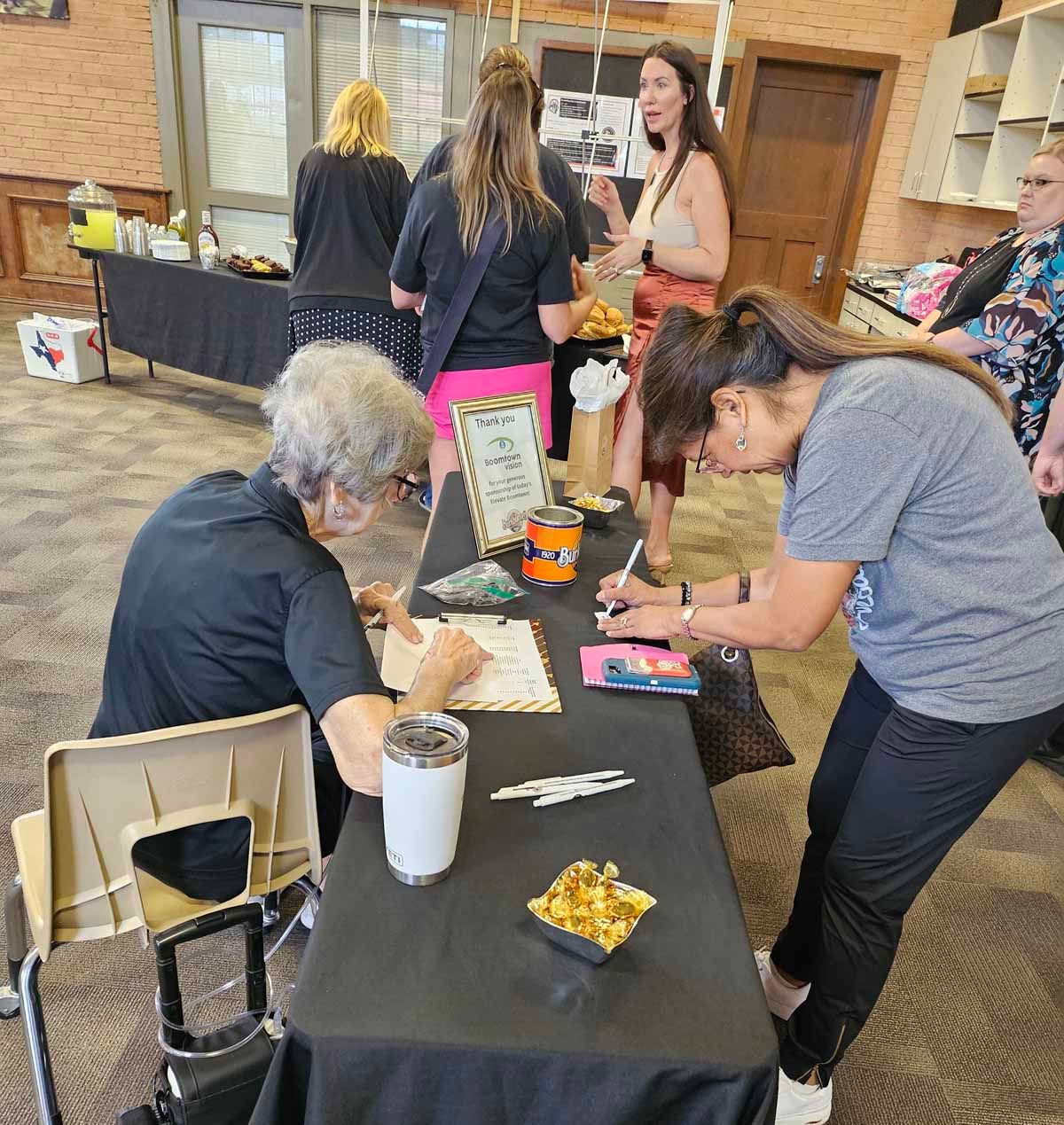 People at a table, some signing documents. Snacks, a drink, and other items sit on the table.