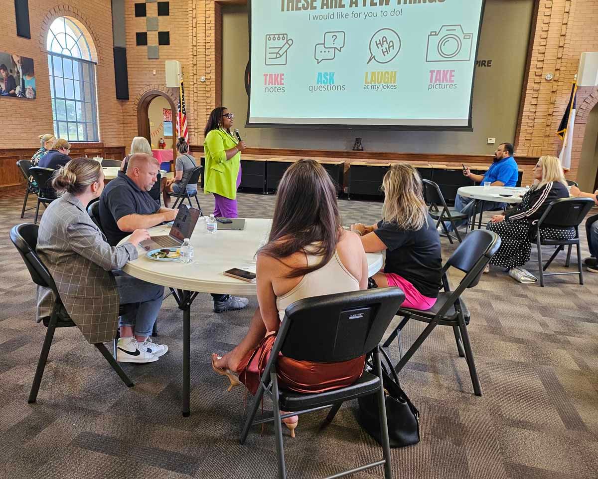 Group of people seated at tables, listening to a presentation on a screen.