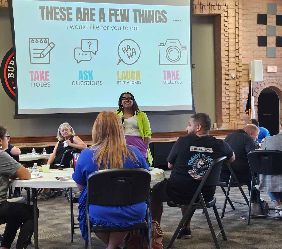 A presenter stands before a seated audience with a projection screen; the screen displays instructions.