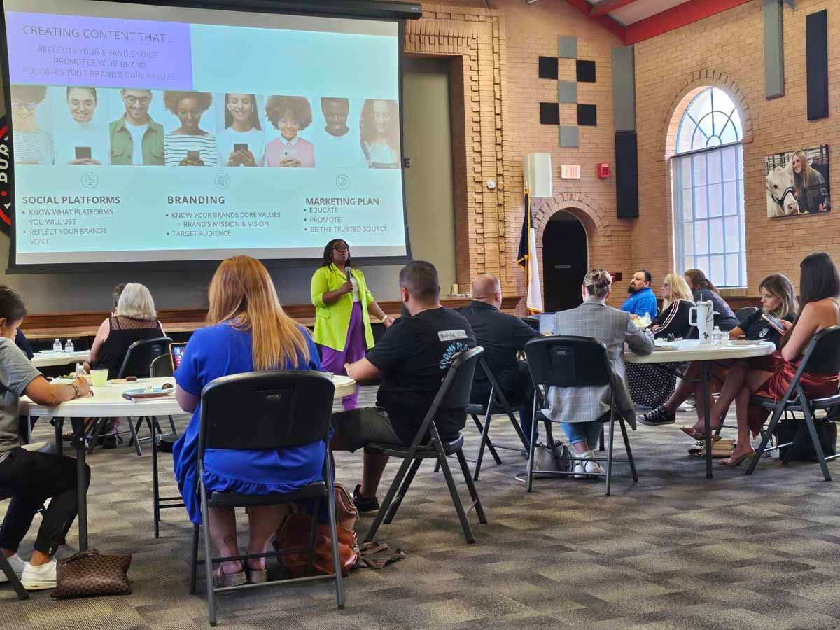 A presenter in a green top speaks at a table event; a projector screen shows faces, and people are seated.