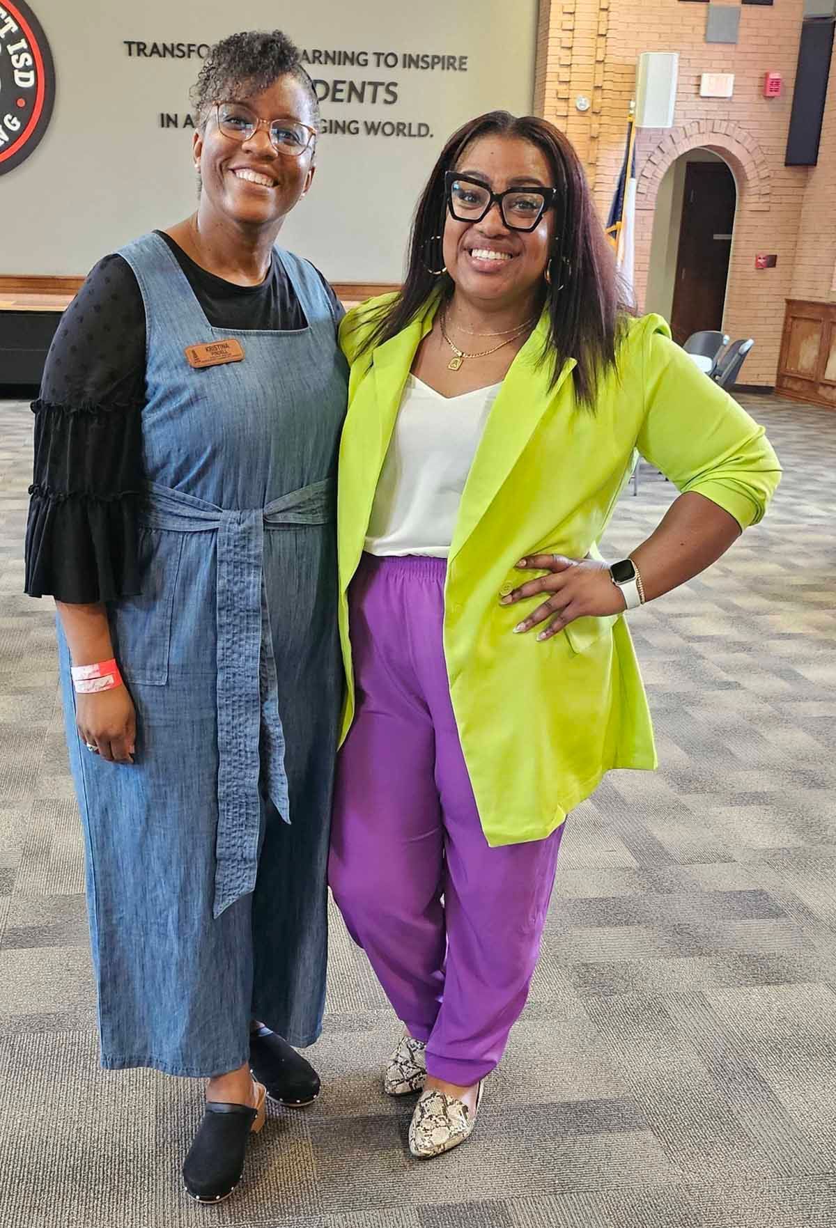 Two women standing together, smiling. One in a denim jumpsuit, the other in purple pants and a green blazer. Indoor setting.