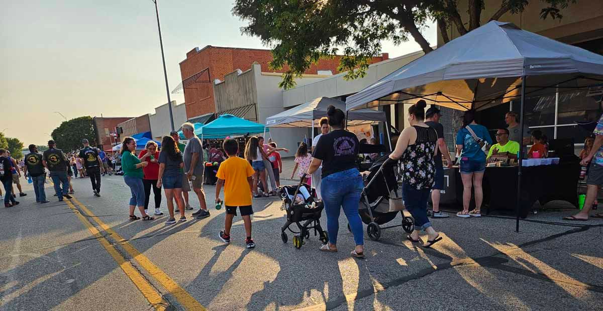 Street festival with people, tents, and buildings. Warm lighting suggests late afternoon.
