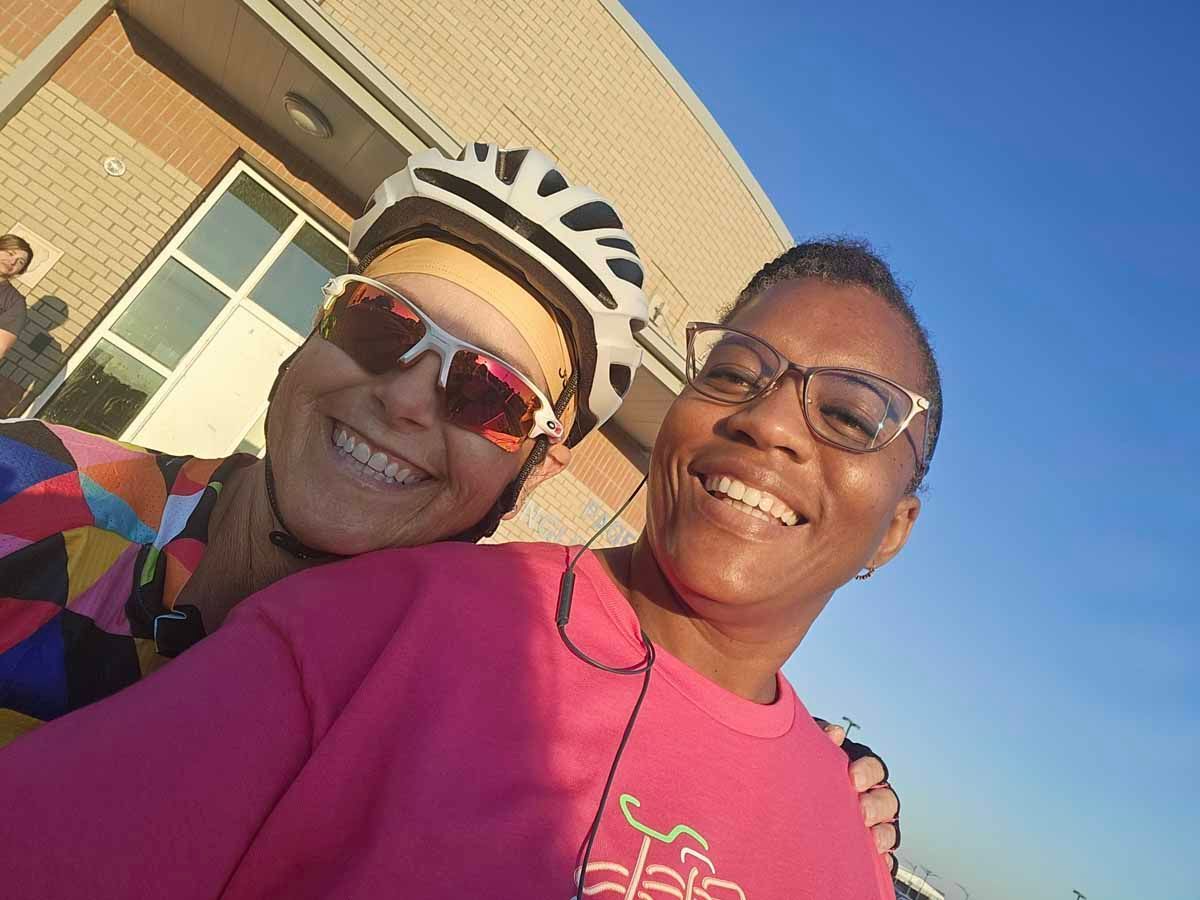 Two women smiling for selfie. One wears a bike helmet and sunglasses, the other has glasses and a pink shirt.