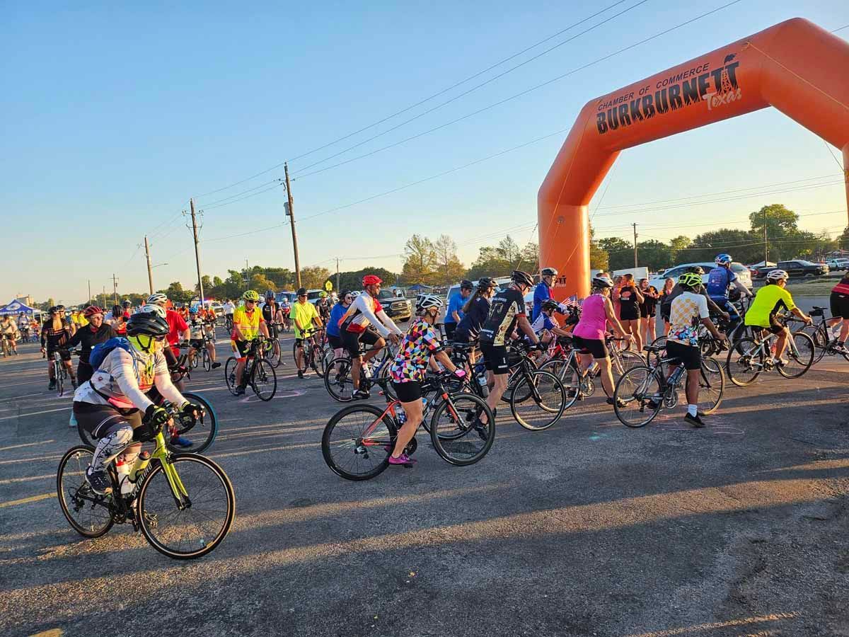 Cyclists lined up at the start of a cycling event under an orange archway.