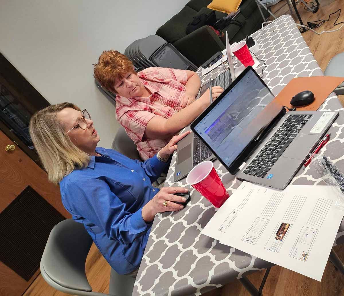 Two people seated at a table with laptops, papers, and red cups; one woman in a blue shirt, the other in a pink plaid shirt.