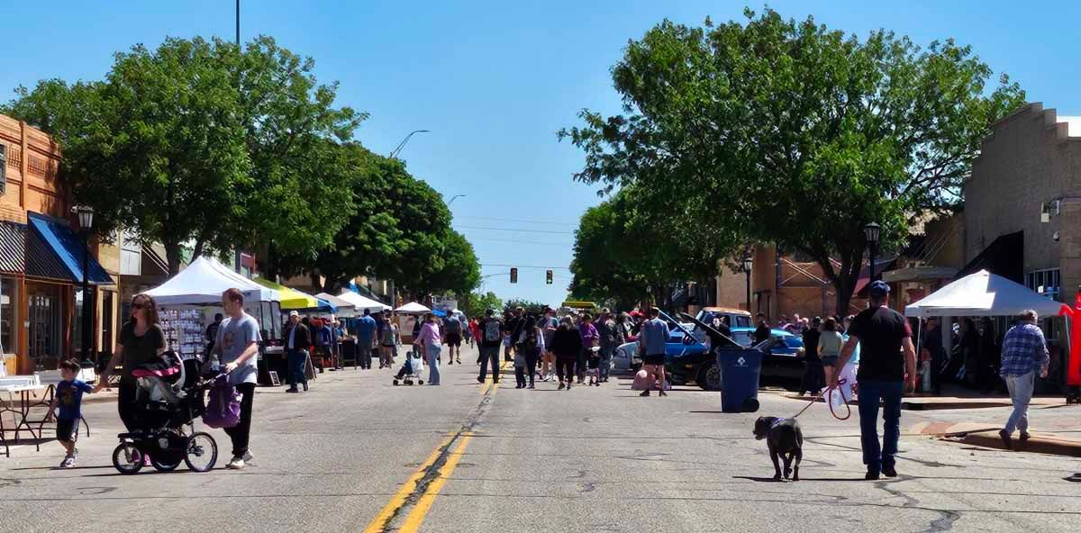 Street fair with pedestrians, tents, and trees on a sunny day. A person walks a dog.