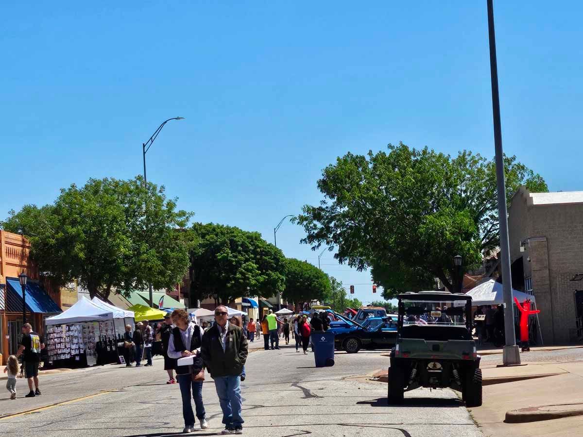 Street fair: people walking on a closed street lined with vendor tents, cars, and trees under a clear blue sky.