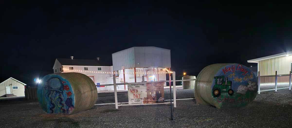 Night scene at a farm with hay bales painted with designs and a structure in between, under a dark sky.