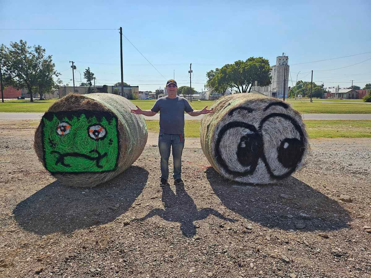 Man stands between two hay bales painted as Frankenstein's monster and a cartoon eye, on a sunny day.