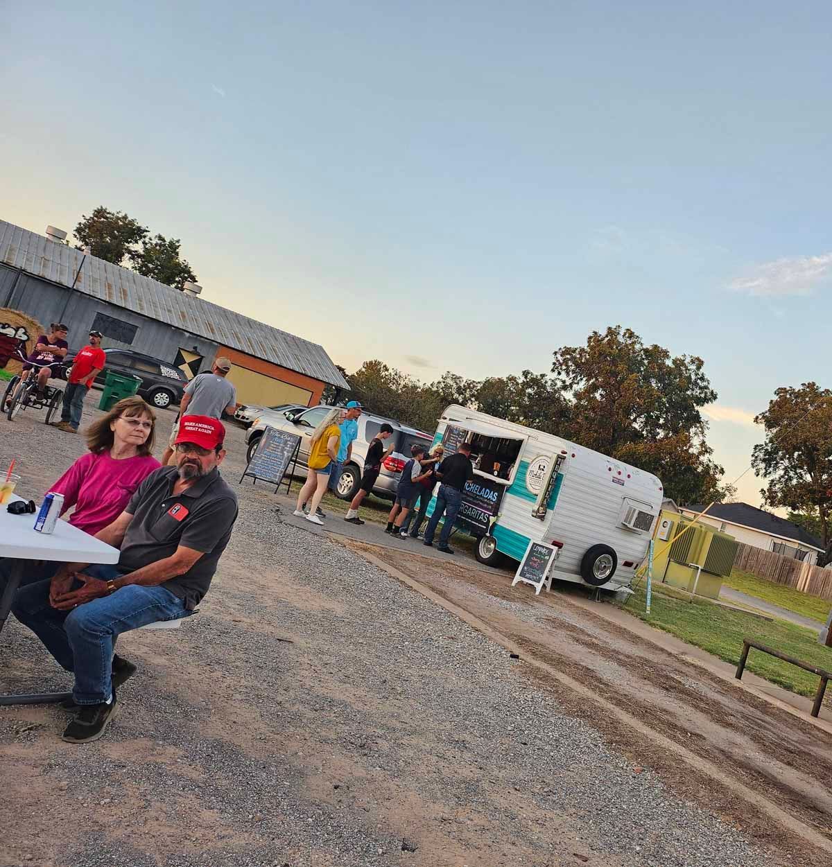 People at an outdoor event with a food truck, seated at tables and socializing. Gravel ground, cloudy sky.
