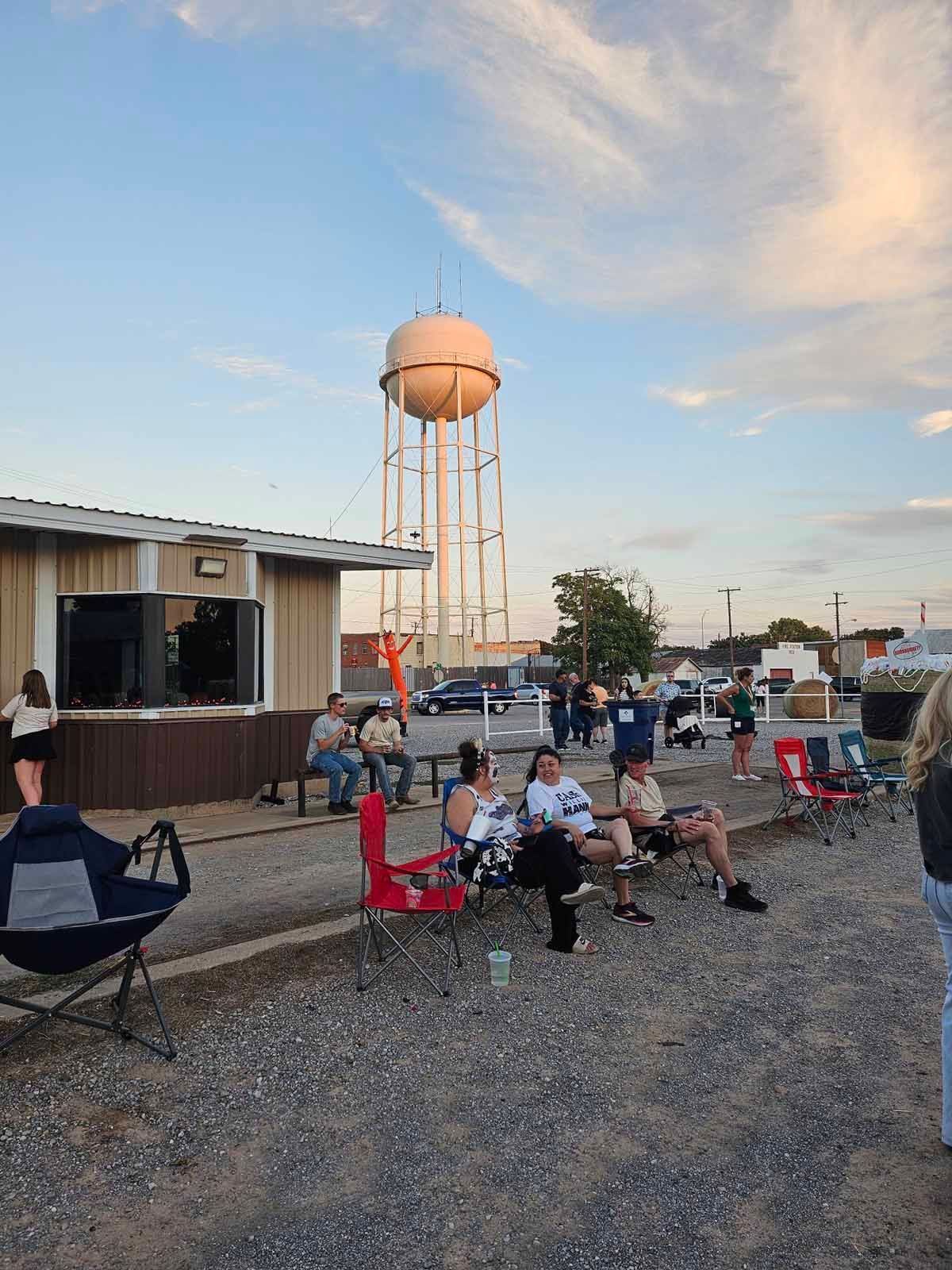 People sitting outside a small building with a water tower in the background, under a partly cloudy sky.