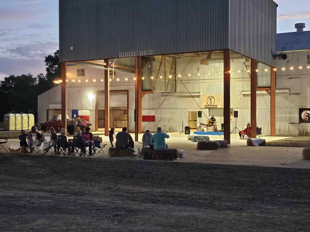 Outdoor music venue: band on stage, audience seated on hay bales, barn setting at dusk.