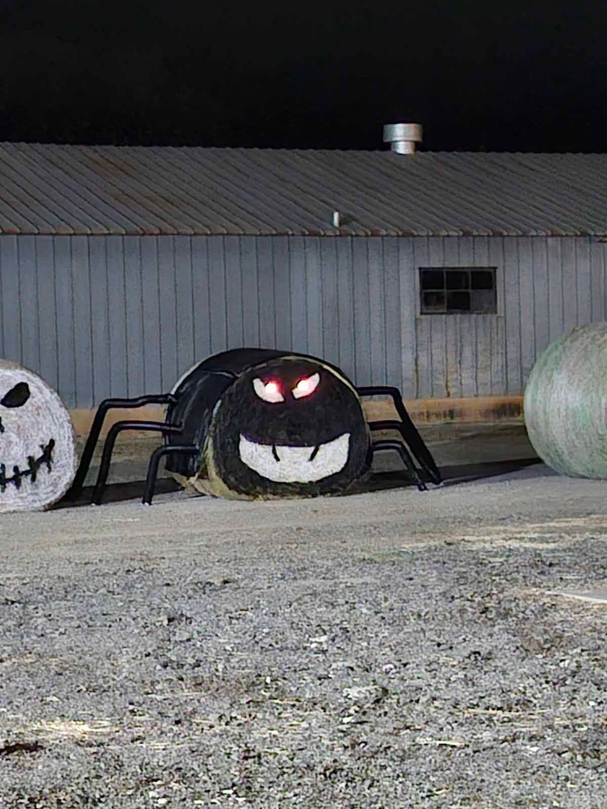 Halloween hay bale decorations: giant spider with red eyes, plus a spooky face and green bale, set before a shed.