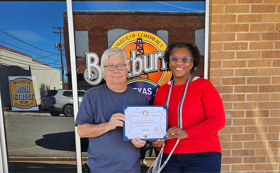 Man and woman holding a certificate outside a brick building. The woman is wearing a red jacket.