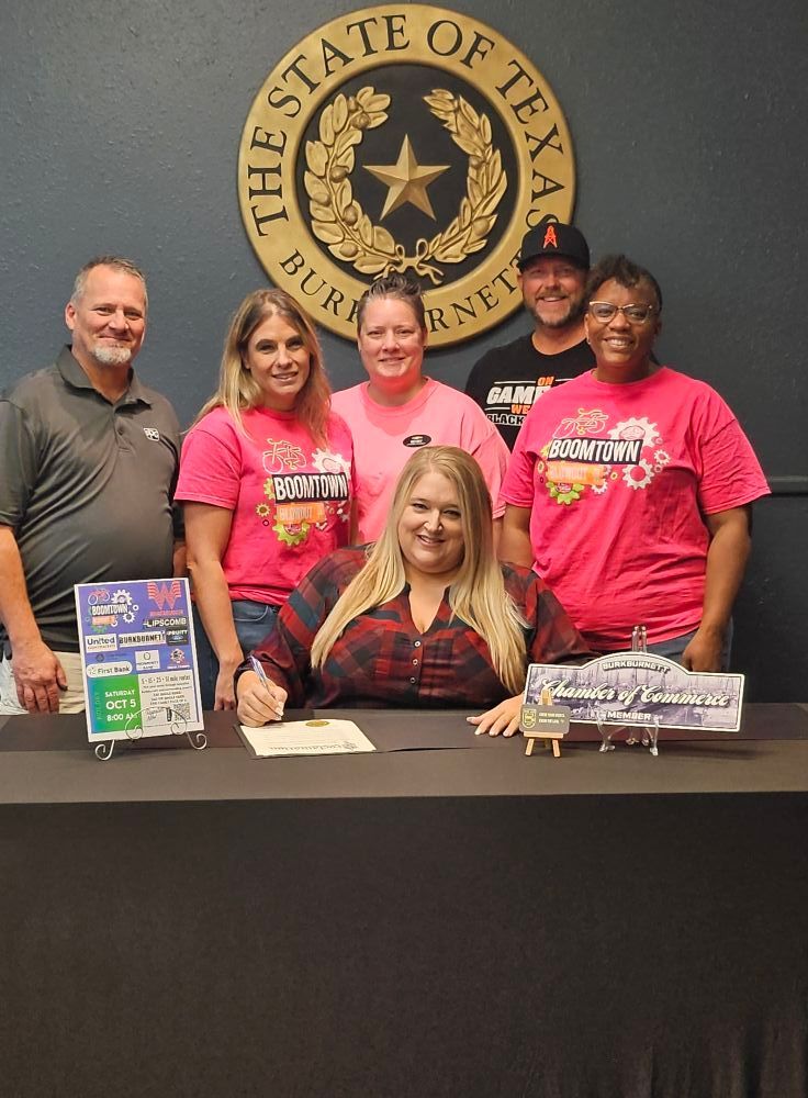 A group signs a document under the Texas state seal; they are wearing pink shirts and smiling.