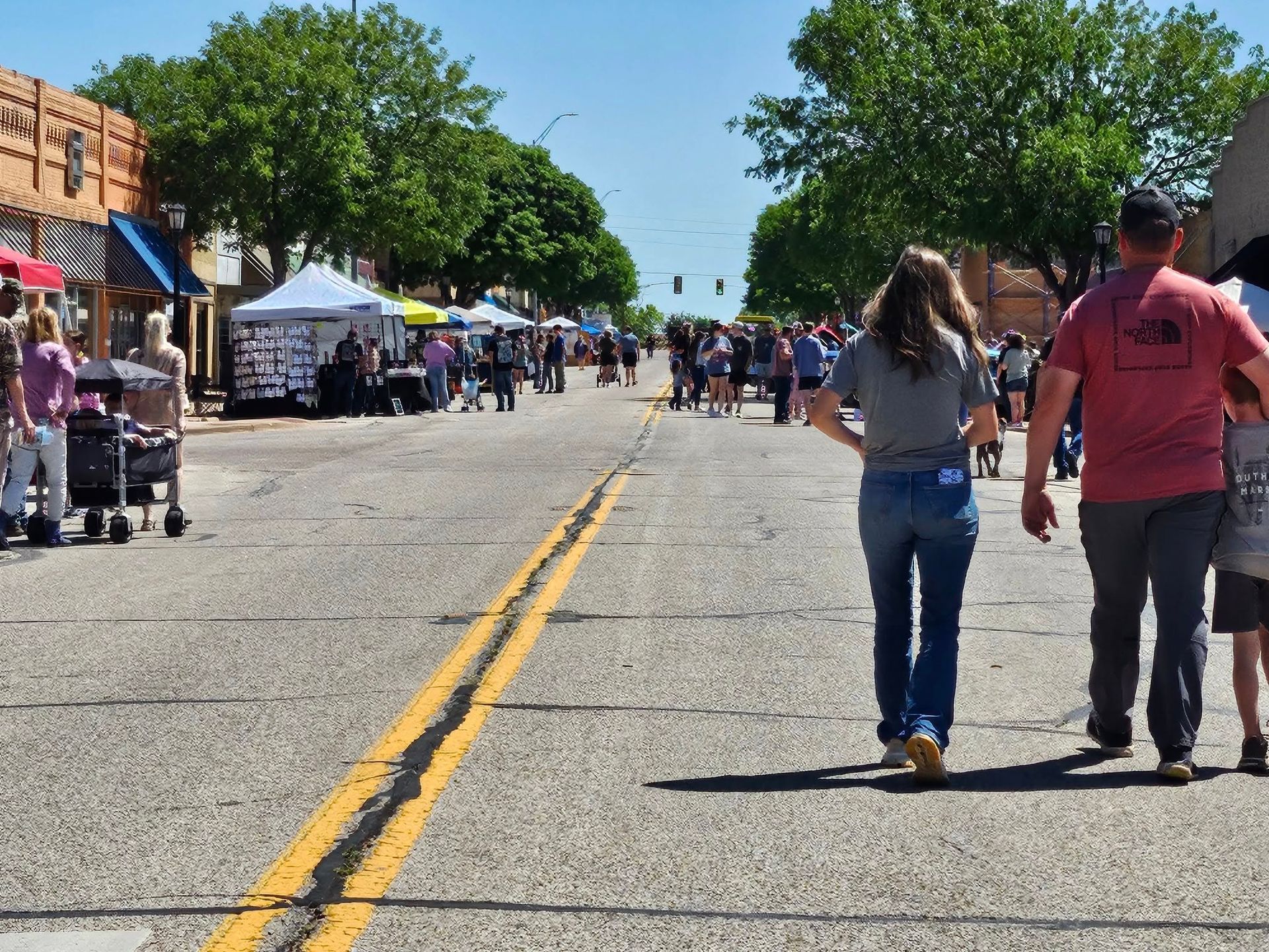 People strolling on a sunny street lined with vendor booths. A family walks towards the viewer.