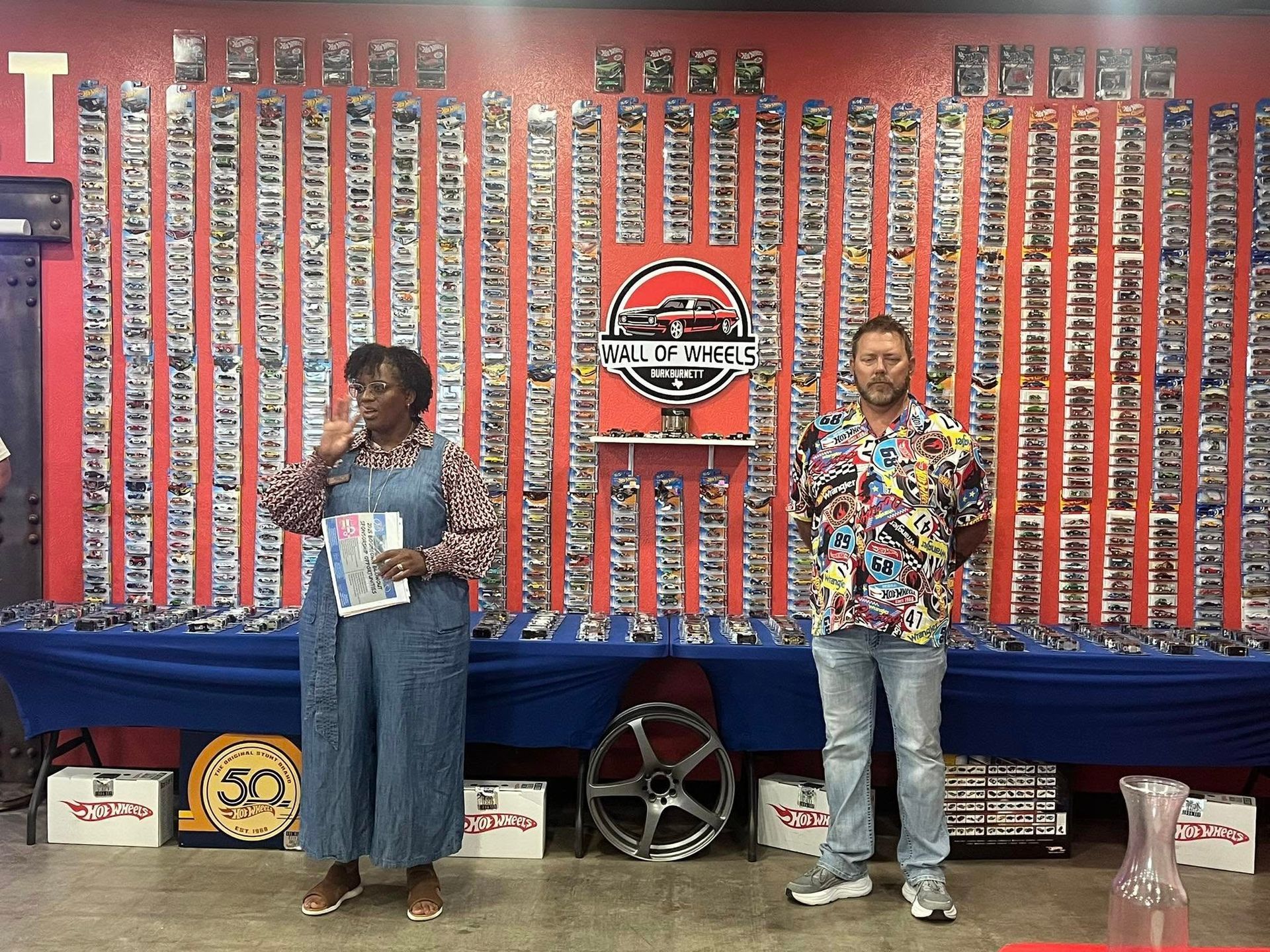 Two people stand before a wall displaying numerous small, collectible items. A woman speaks, holding papers.
