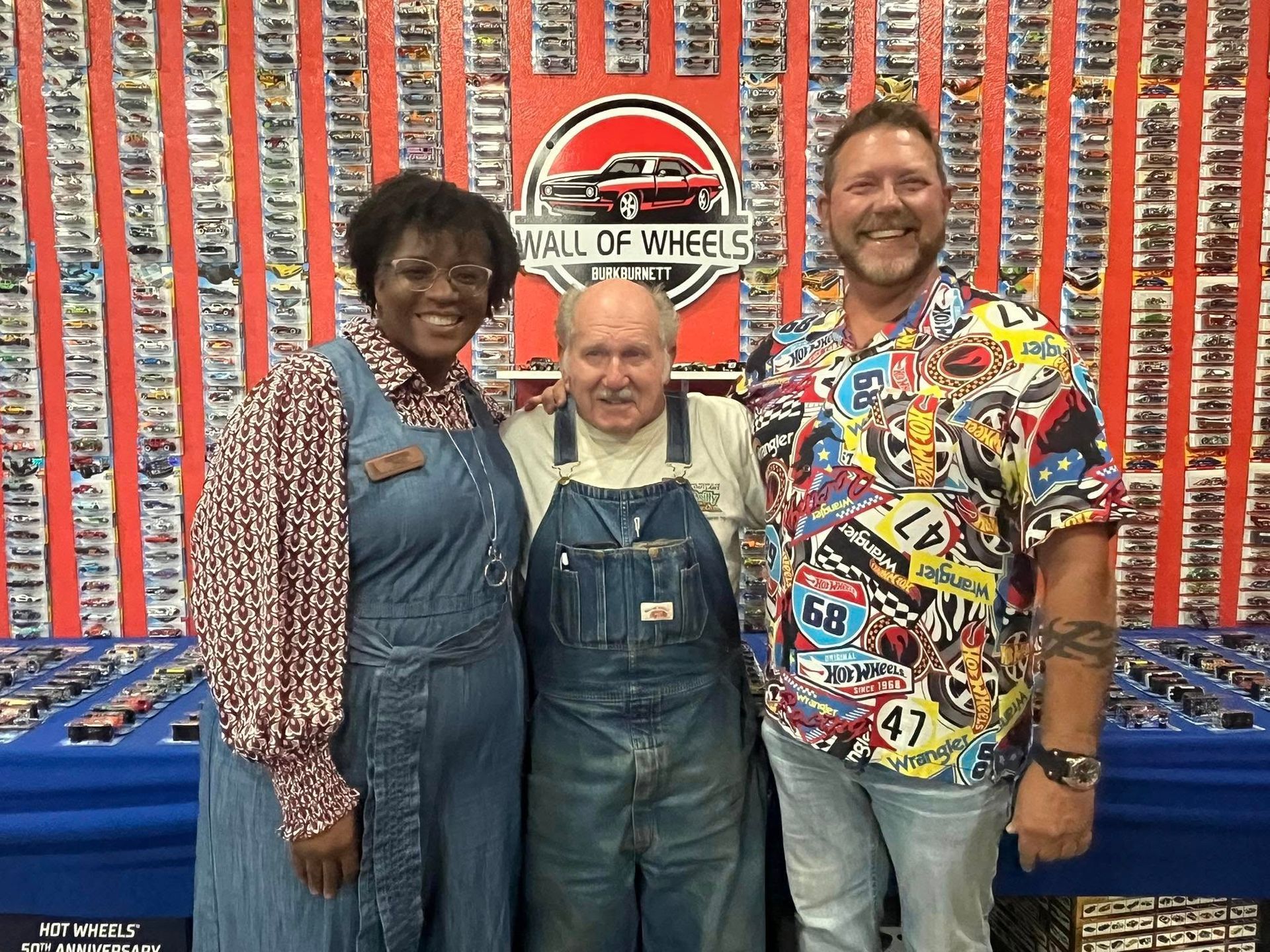 Three people posing in front of a wall of toy cars. The middle man wears denim overalls, the others wear colorful tops.