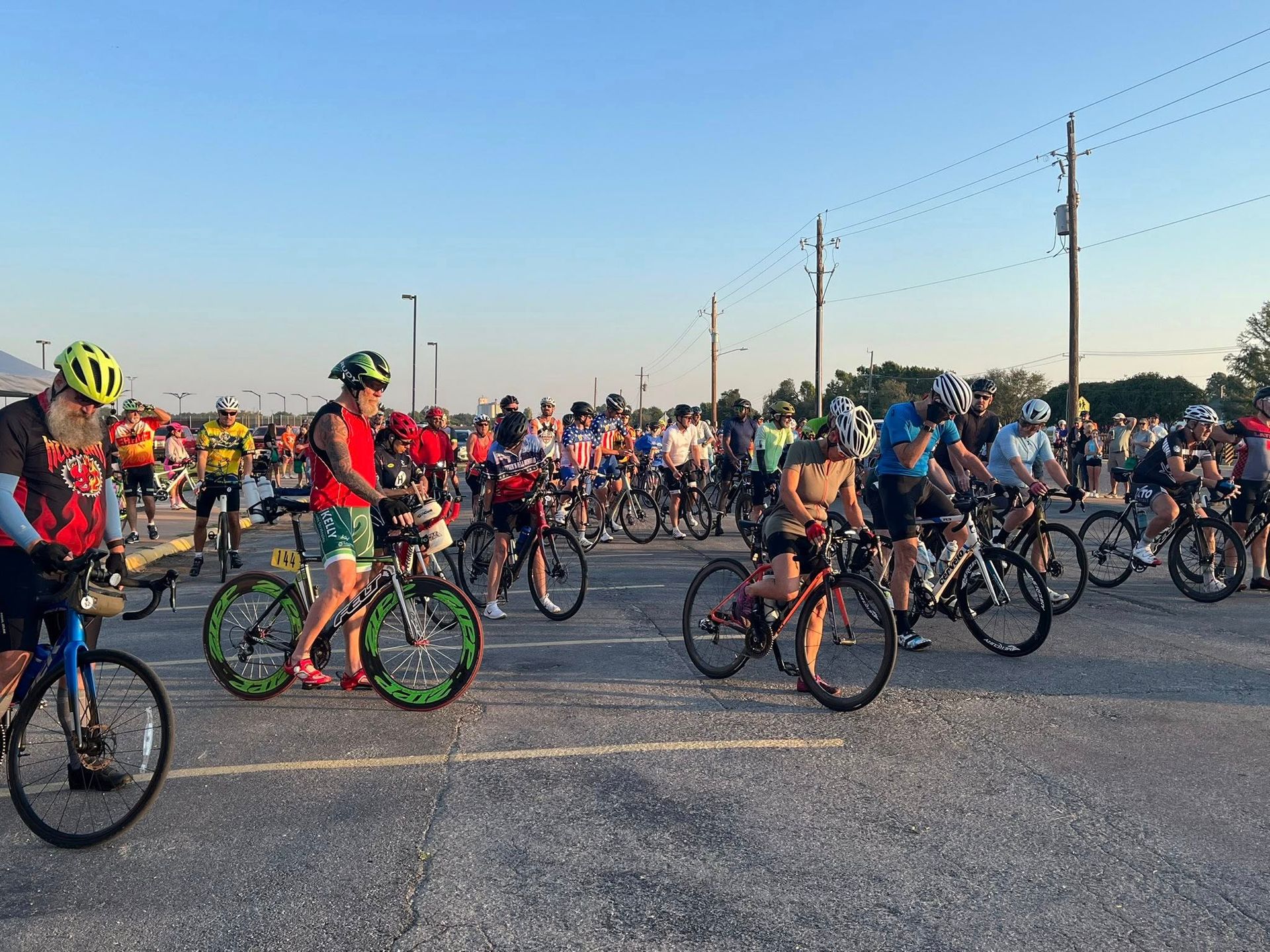 Cyclists on bikes, wearing helmets, gathered on a paved road, ready to start a ride on a sunny day.