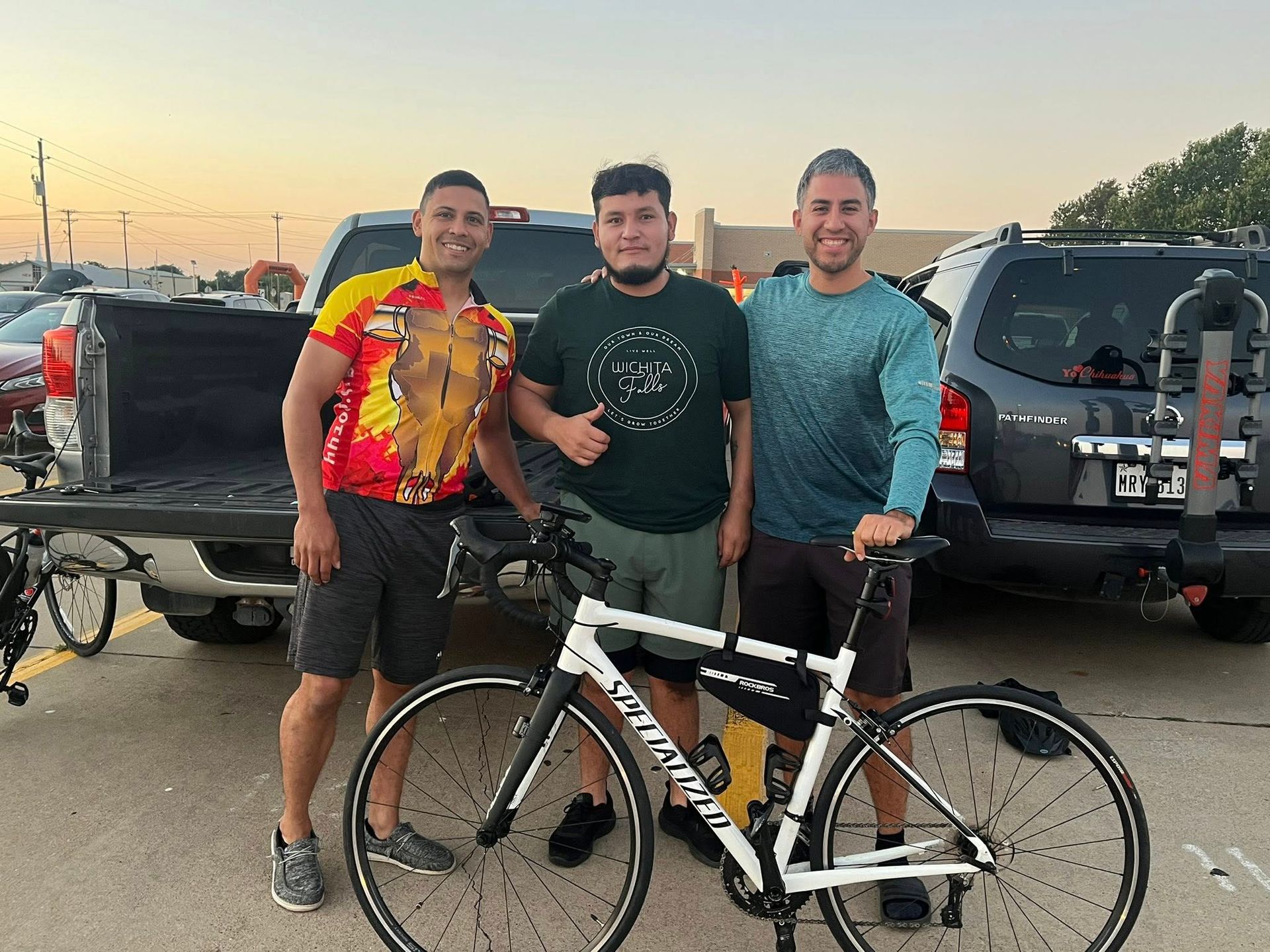 Three people pose with a white Specialized bicycle in a parking lot. Two men stand on either side of the man holding the bike.