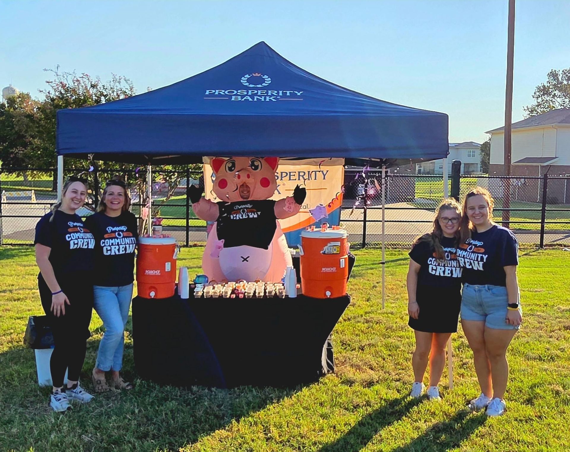 People at a booth with a pig mascot. They are standing under a tent. There are orange coolers and drinks.