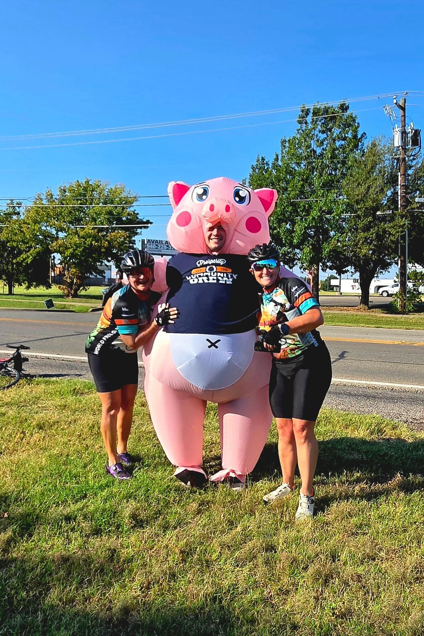 Two cyclists flank a person in a large inflatable pig costume on a grassy area, sunny day.