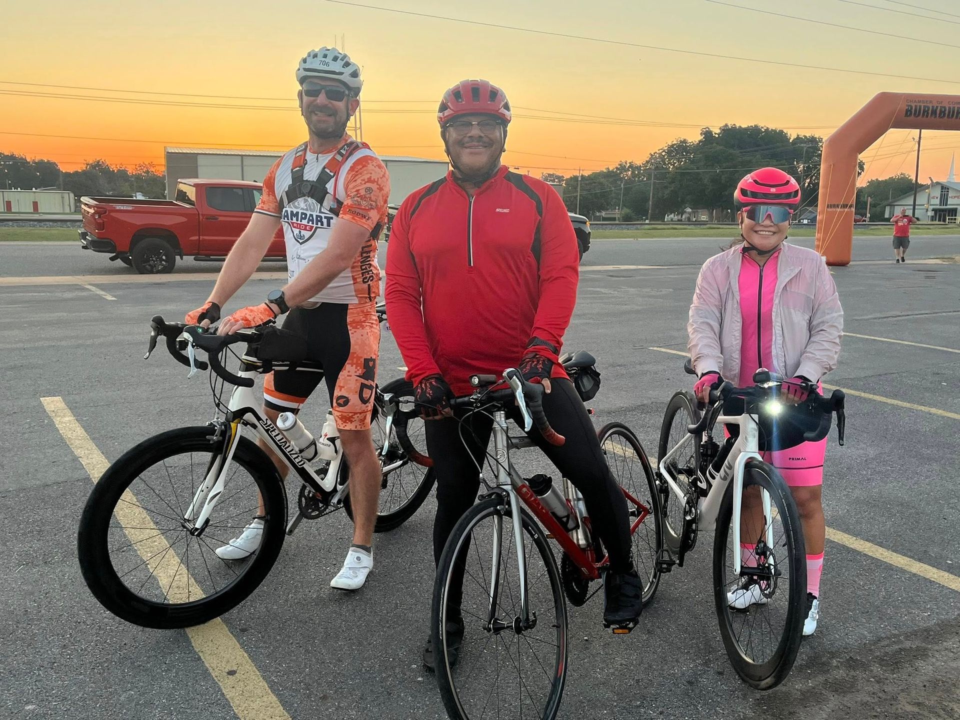 Three cyclists pose with their bikes, outdoors at dusk; orange arch and truck in the background.
