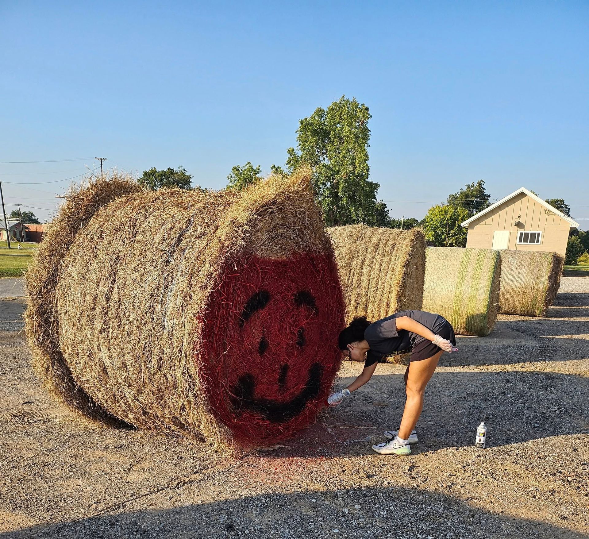 Woman spray-painting a large hay bale to resemble the Kool-Aid Man. The setting is outdoors on a sunny day.