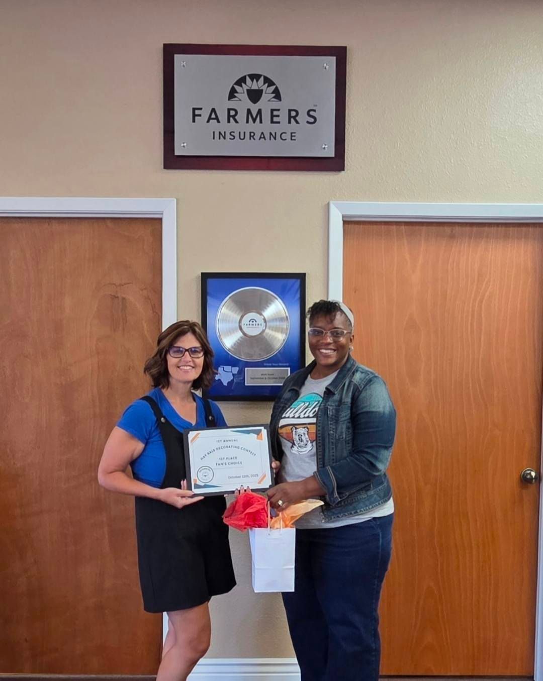 Two people holding award in front of Farmers Insurance sign. One person wears a denim jacket and blue jeans.