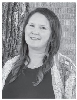 Woman smiling outdoors, leaning against a tree trunk and brick wall, wearing a patterned cardigan.