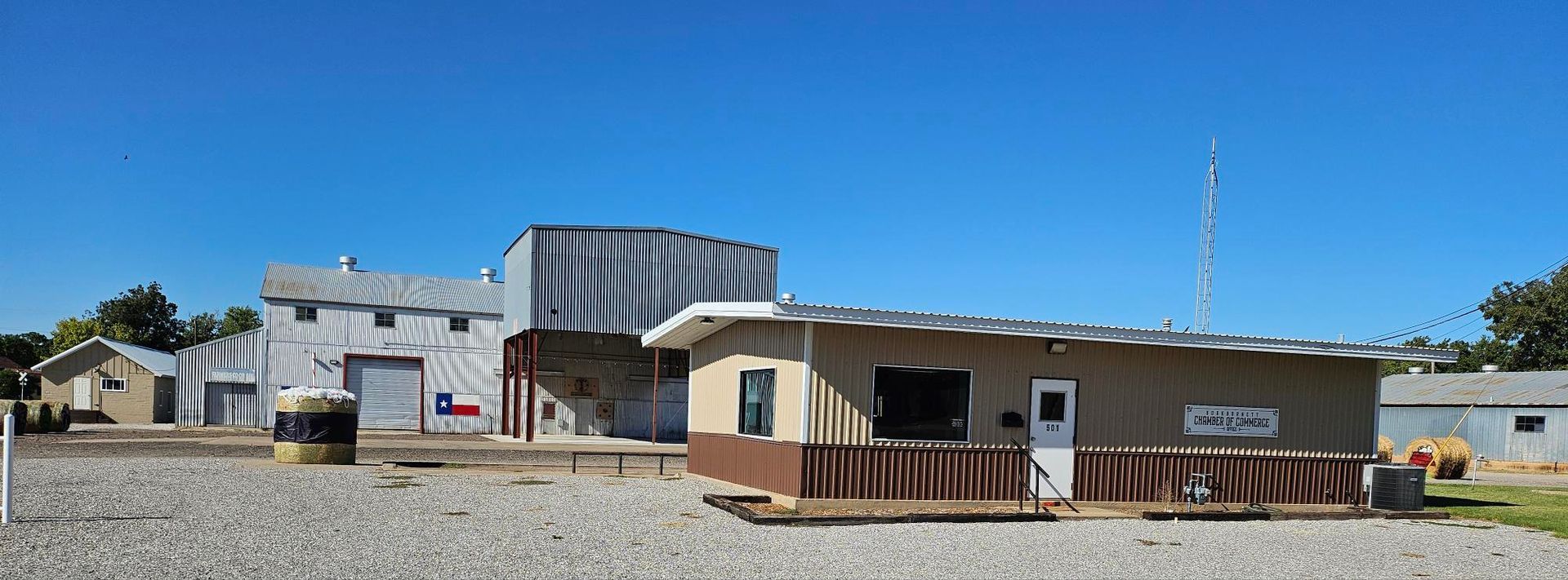 Buildings with metal roofs and Texas flag. Bright blue sky. Gravel lot.