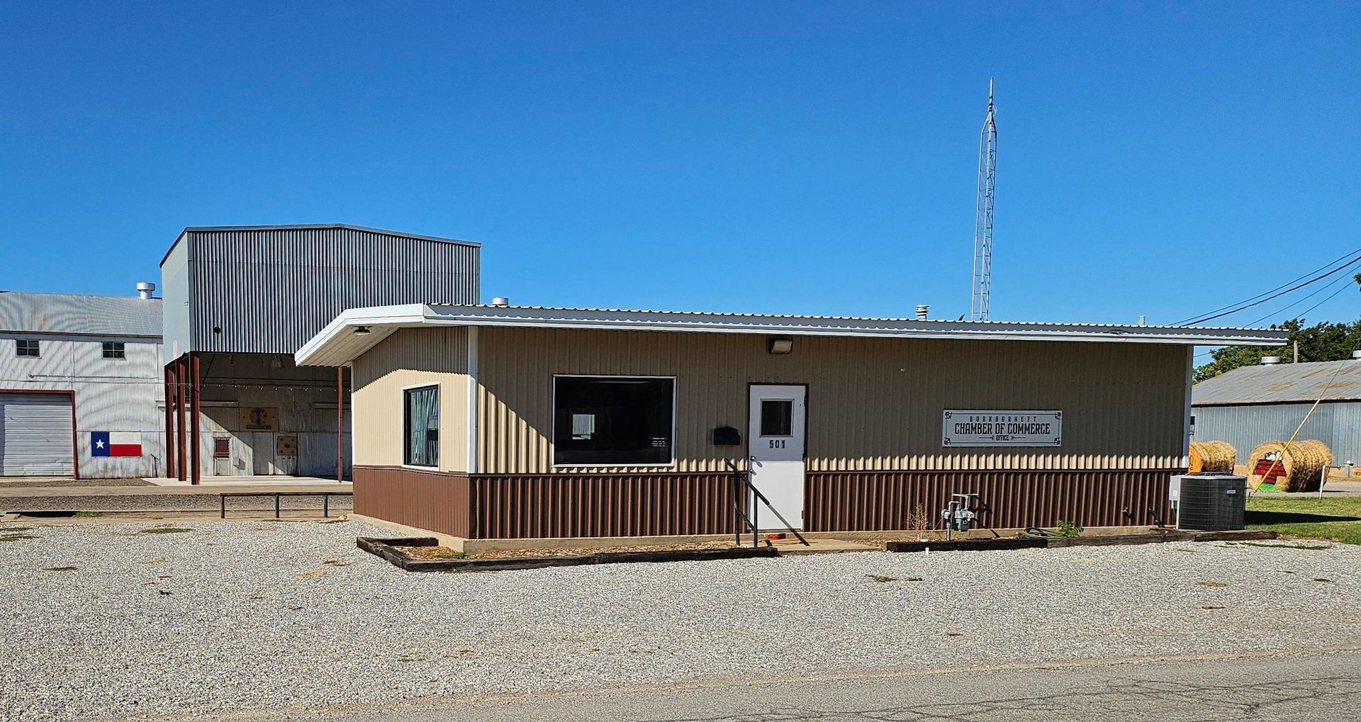Small building with sign, brown and tan, next to industrial building and gravel lot under blue sky.