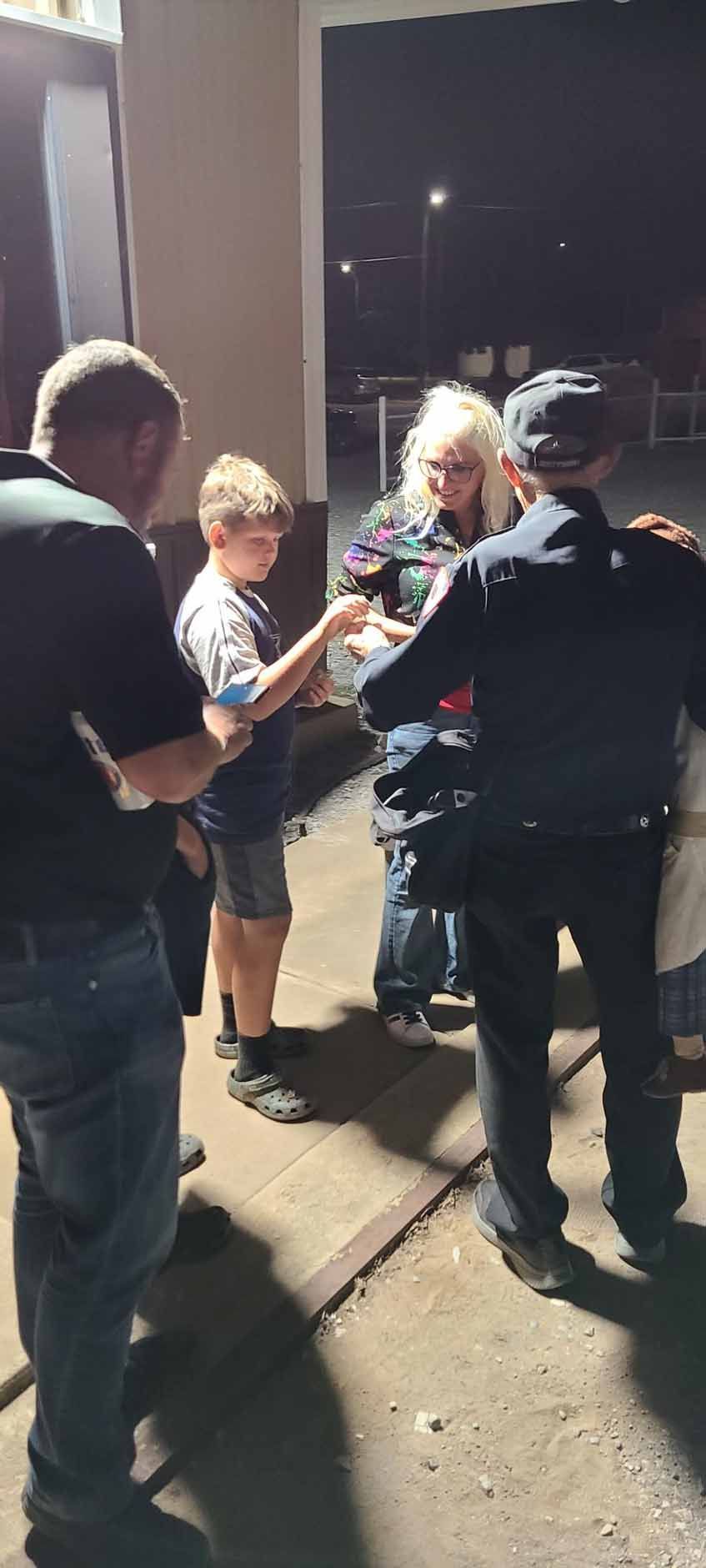 People standing in front of a building at night. A young person hands something to a blonde person.