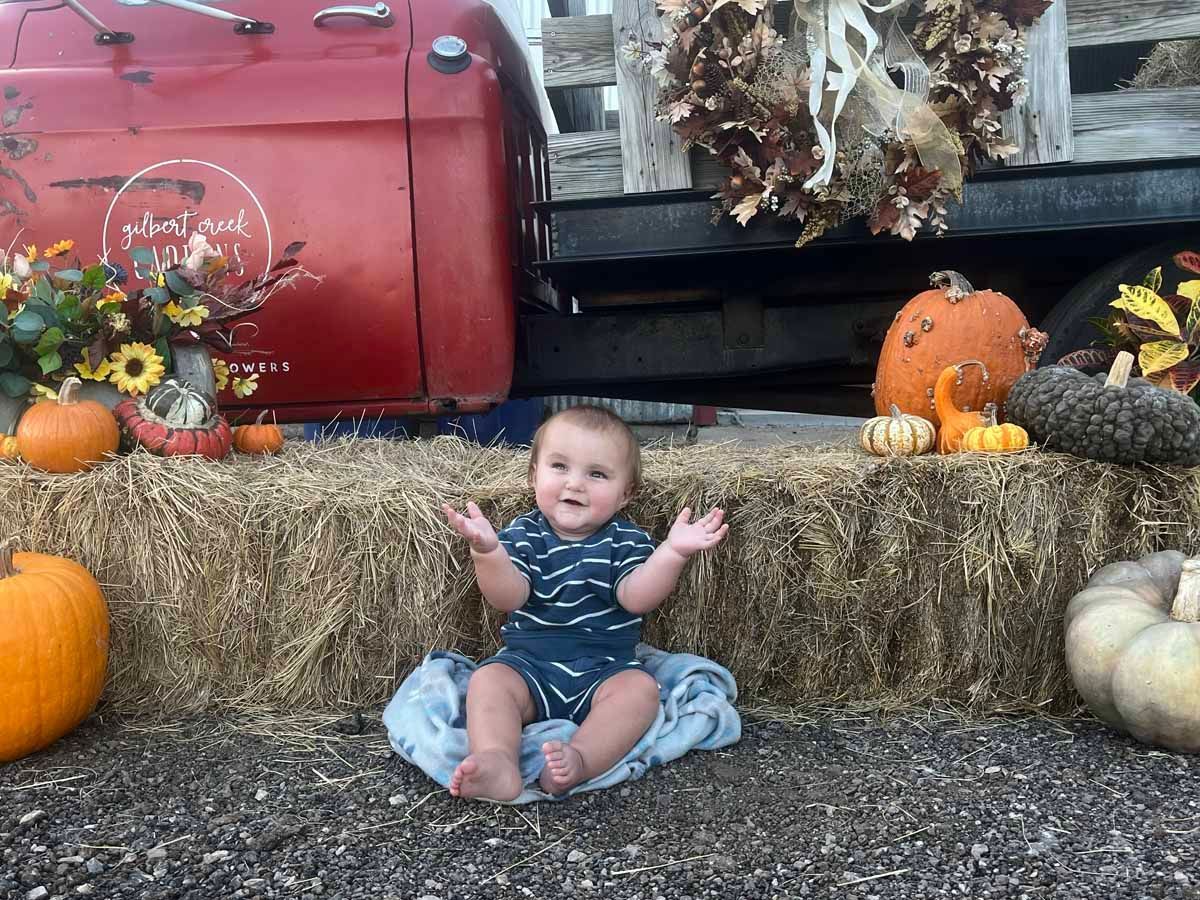 Baby sits with arms raised in front of hay bales, pumpkins, and a red truck bed decorated for fall.