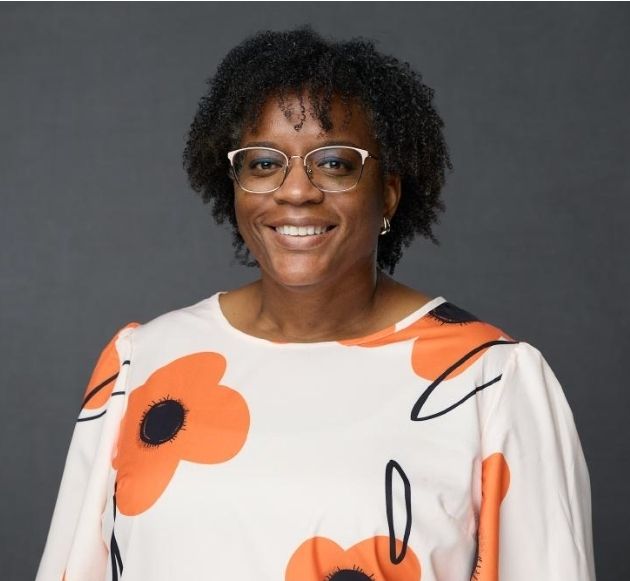 Woman with glasses smiles, wearing floral top, set against gray background.