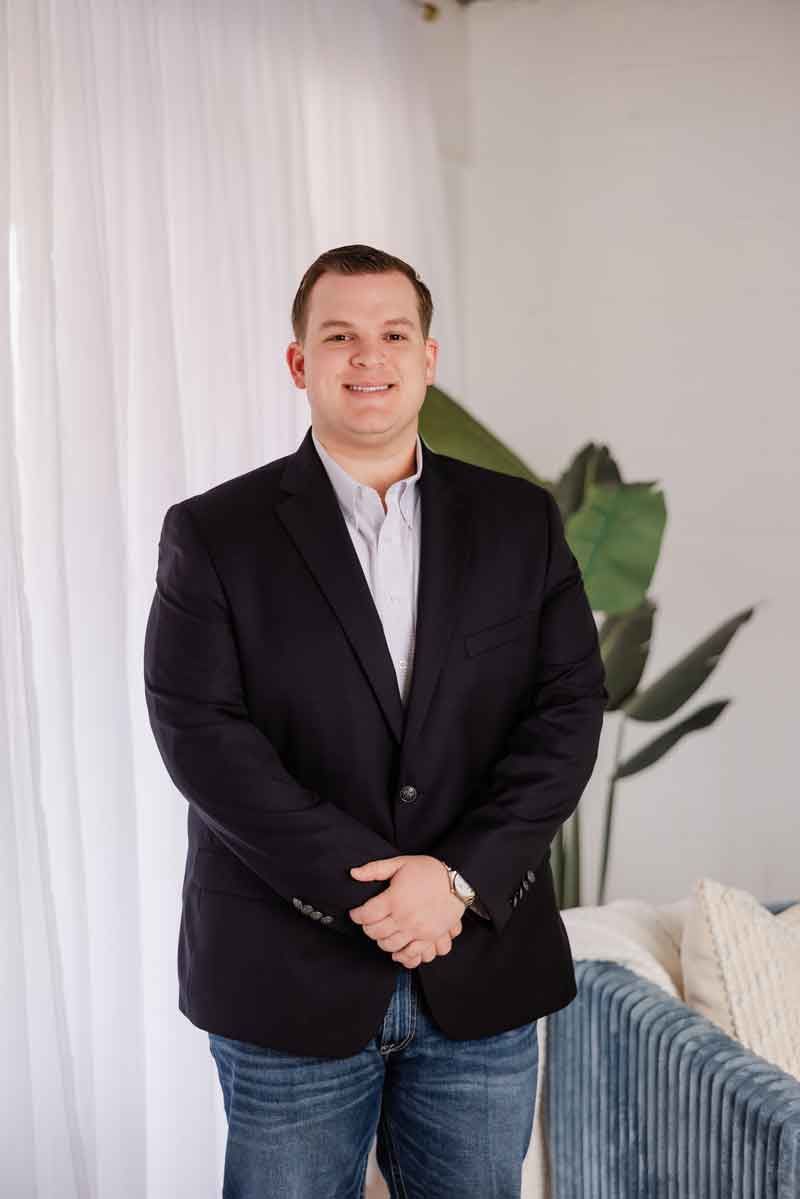 Man in navy blazer and jeans, hands clasped, smiling slightly. Standing indoors near a sofa.