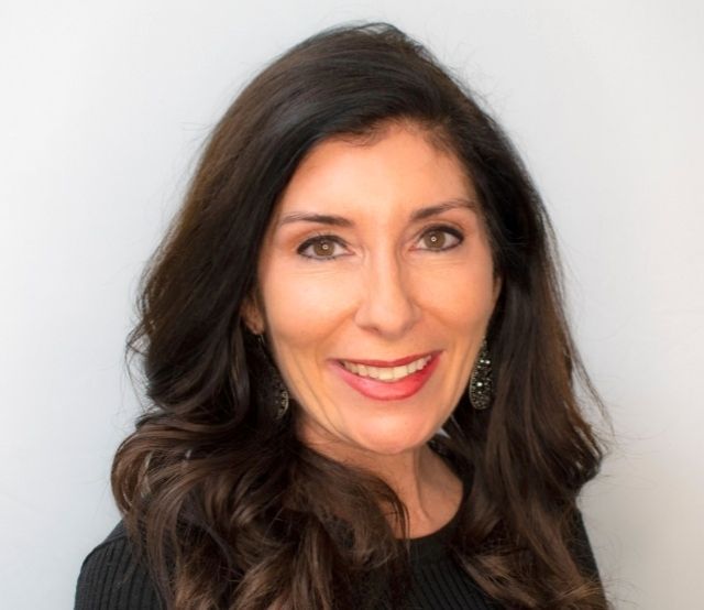 Woman with long, dark wavy hair, smiling and wearing a black top against a light gray background.