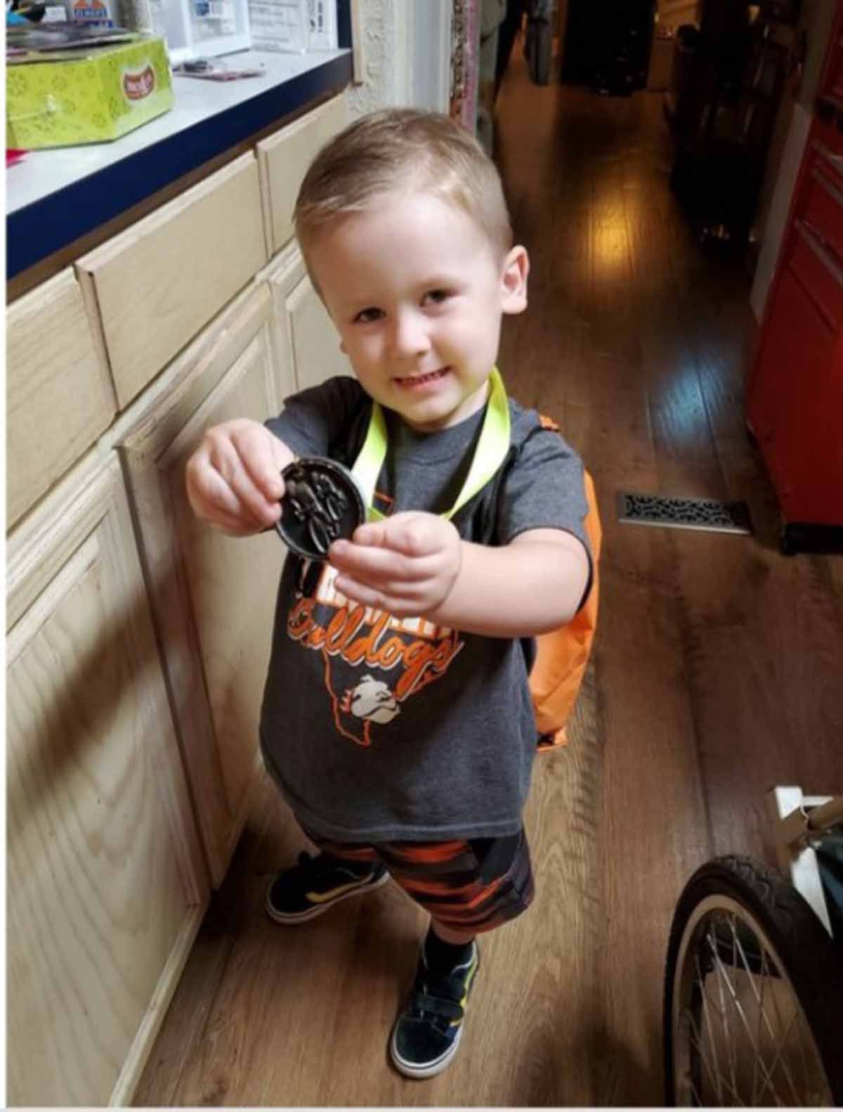 Smiling child holding a medal, wearing a backpack and athletic wear, standing indoors.