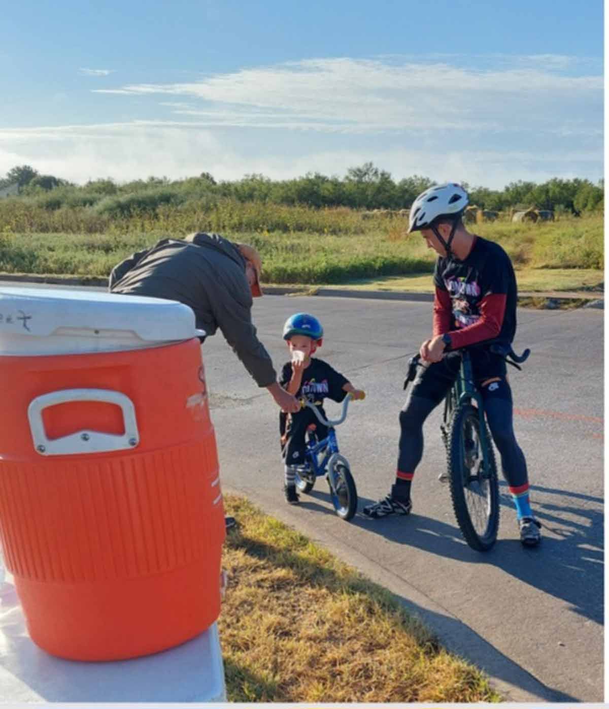 Man giving drink to child on balance bike, cyclist watches, next to an orange cooler outdoors.