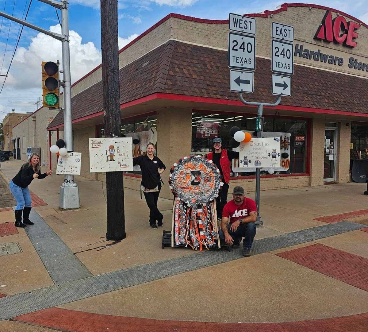 People pose with a large, elaborate mum outside an Ace Hardware store, street signs in the background.