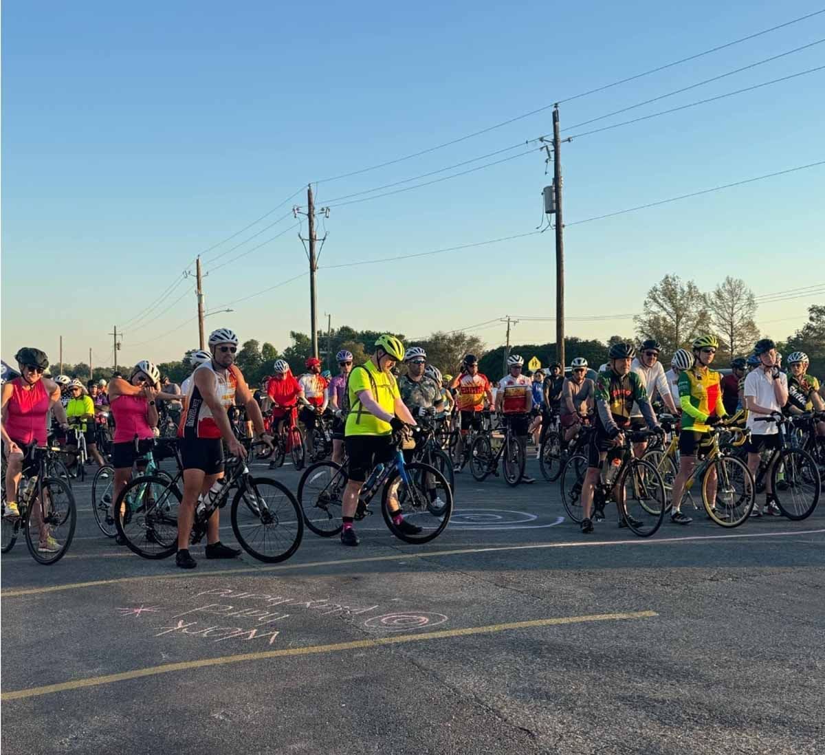 Cyclists on bikes, lined up on a road, preparing to start a ride. Dusk setting, power poles in the background.