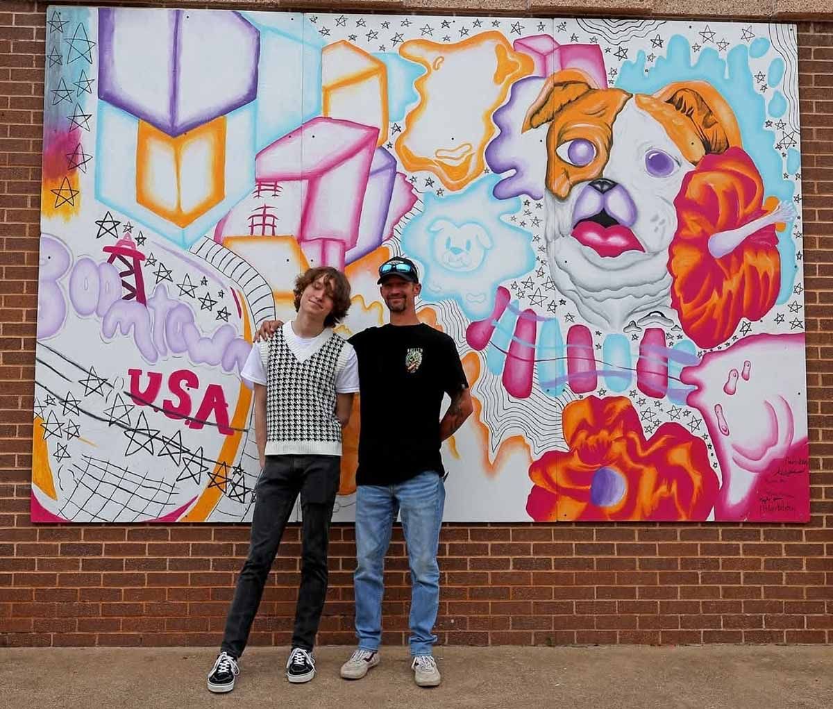 Two people stand in front of a colorful mural featuring a dog and geometric shapes. Brick wall setting.
