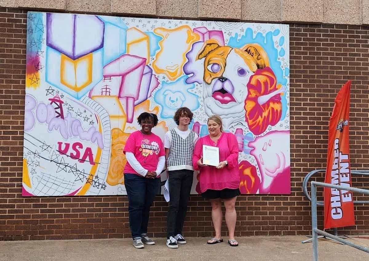 Three people stand in front of a colorful mural of a dog and abstract shapes. They are in front of a brick building.