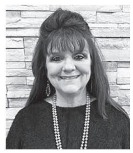 Woman smiling, wearing a pearl necklace, standing in front of a textured wall.