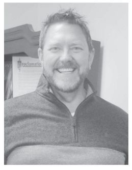 Man smiling, wearing a gray zip-up sweater. Background includes a document and a cabinet.